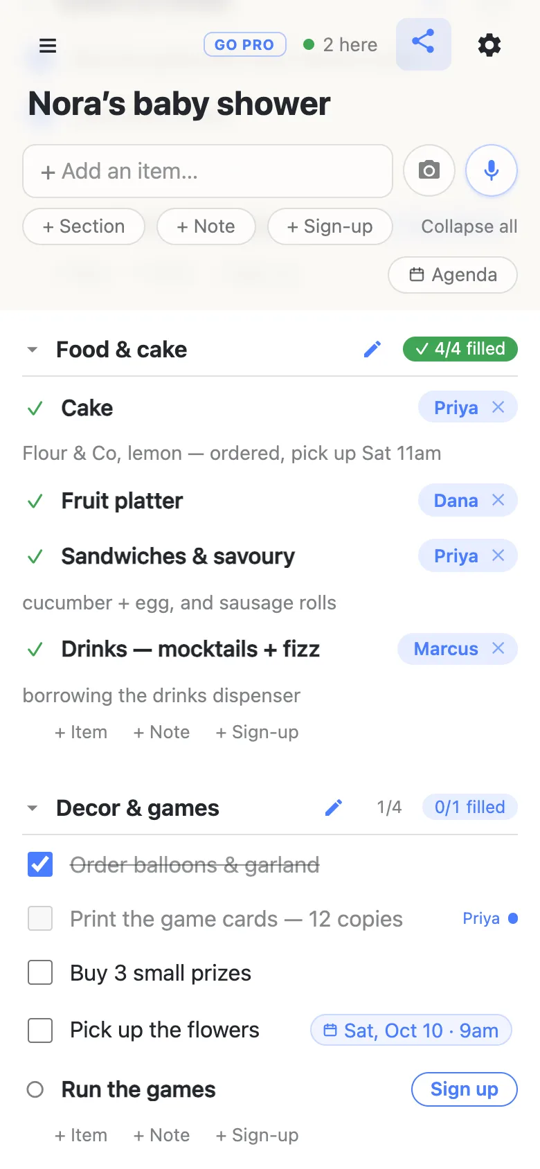 The same list on Dana’s phone a moment later. The header chip reads “2 here” with a green dot. Food & cake now reads a green 4/4 filled: Sandwiches & savoury is claimed by Priya with the note “cucumber + egg, and sausage rolls”. In Decor & games the row “Print the game cards — 12 copies” is greyed with the name Priya and a dot beside it, because Priya is editing it right now.