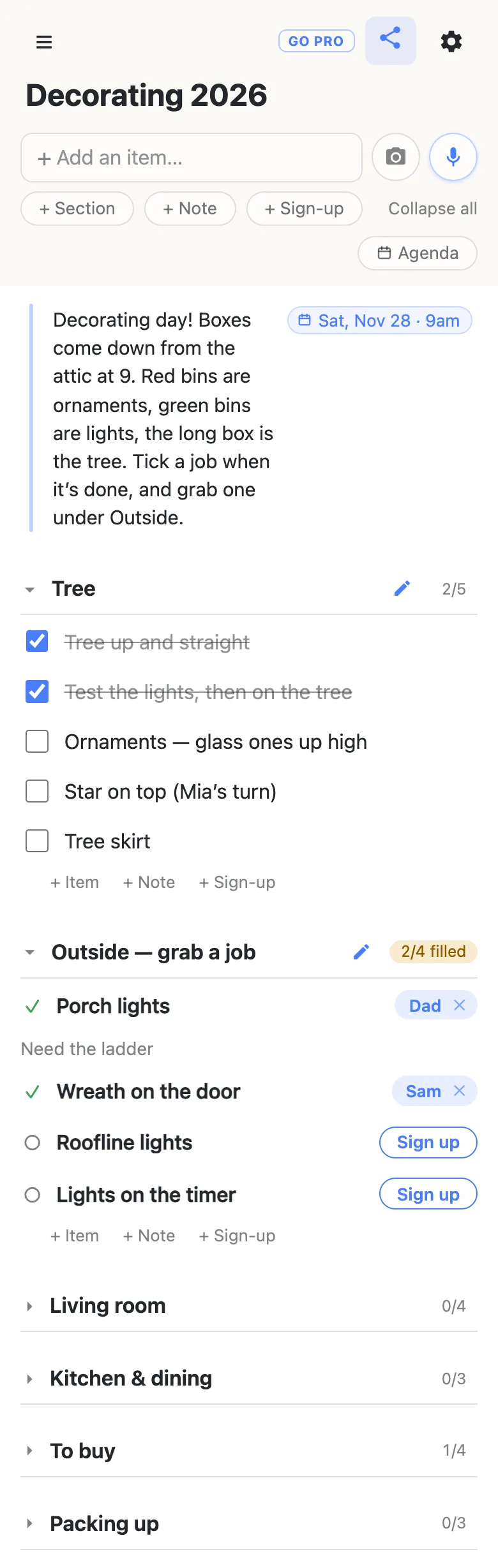 A phone showing a list titled “Decorating 2026”. A note at the top, with a date chip reading Sat, Nov 28 · 9am, says: Decorating day! Boxes come down from the attic at 9. Red bins are ornaments, green bins are lights, the long box is the tree. Tick a job when it’s done, and grab one under Outside. The Tree section reads 2/5: Tree up and straight and Test the lights, then on the tree are ticked and struck through; Ornaments — glass ones up high, Star on top (Mia’s turn) and Tree skirt are open. Outside — grab a job reads 2/4 filled: Dad has the Porch lights with the note Need the ladder, Sam has the Wreath on the door, and Roofline lights and Lights on the timer each show a Sign up button. Living room (0/4), Kitchen & dining (0/3), To buy (1/4) and Packing up (0/3) are folded shut underneath.