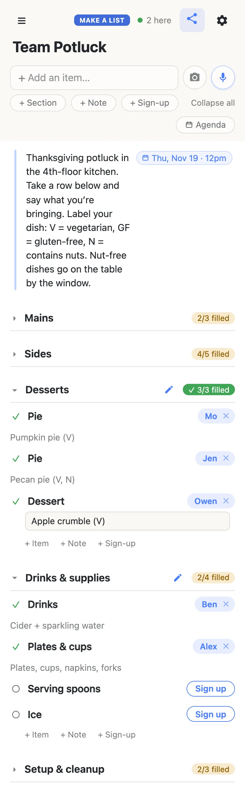 A coworker’s phone showing the same Team Potluck list, with “2 here” and a green dot in the header. The note and its Thu, Nov 19 · 12pm chip sit at the top; Mains (2/3 filled) and Sides (4/5 filled) are folded shut. Desserts is open and now reads 3/3 filled in green with a tick: Mo has a Pie (“Pumpkin pie (V)”), Jen a Pie (“Pecan pie (V, N)”), and Owen has just signed up for Dessert and is typing “Apple crumble (V)” into the note box beneath it. Drinks & supplies reads 2/4 filled: Ben has Drinks (“Cider + sparkling water”), Alex has Plates & cups (“Plates, cups, napkins, forks”), and Serving spoons and Ice show Sign up buttons. Setup & cleanup is folded at 2/3.